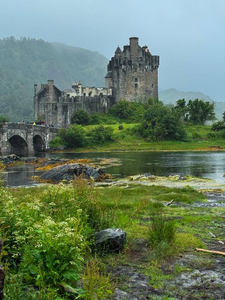 Iconic Eilean Donan Castle and stone bridge reflected in tidal waters with misty highlands behind.