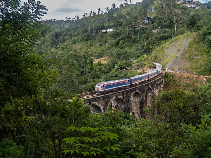 Blue and red train crosses the historic Nine Arches Bridge amid lush green jungle near Ella.