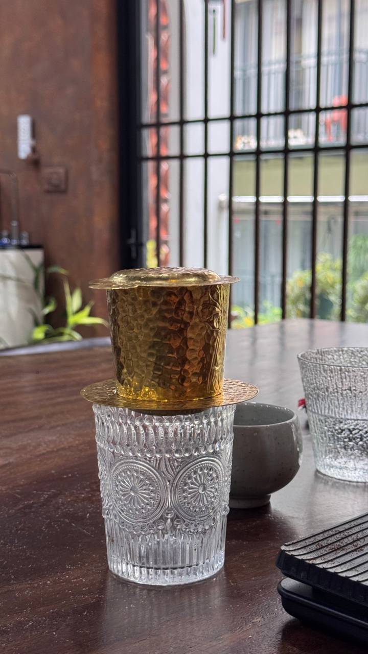 Close-up of a hammered brass Vietnamese coffee filter resting on textured glass cups indoors