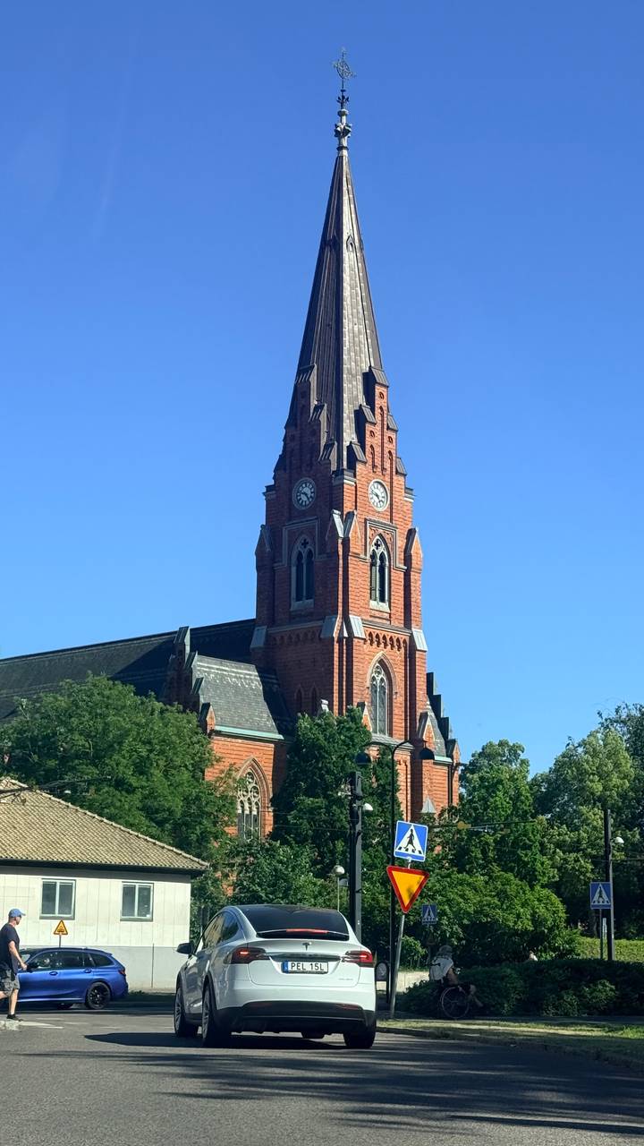 Tall red-brick neo-gothic church tower with clock faces rising above green trees and a vivid blue sky.