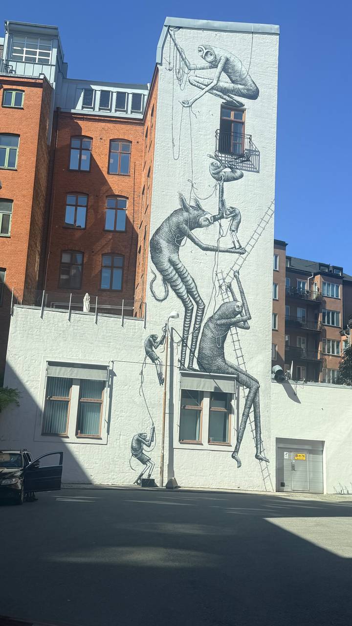 Large monochrome street-art mural depicting elongated figures climbing a wall on an urban building facade.