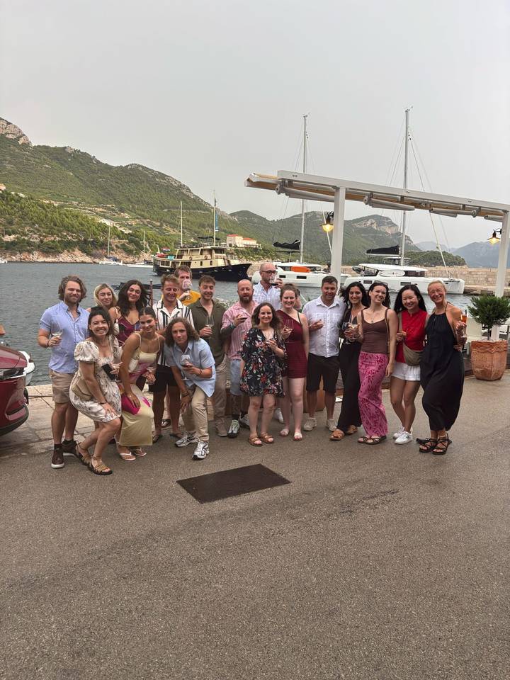 Well-dressed travelers toast with wine by a marina lined with yachts and green hills.