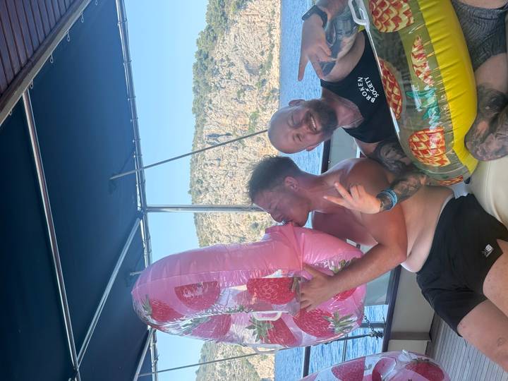 Two men on a boat laugh while holding inflatable pool floats against a rocky island backdrop.