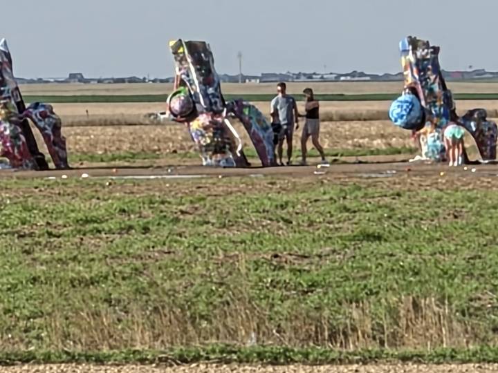 Visitors examine a brightly painted, half-buried car installation in a flat prairie field along Route 66.