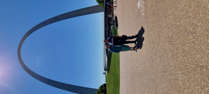 Couple poses beneath the towering steel Gateway Arch against a clear blue sky.