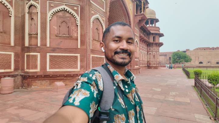 Smiling traveller takes a selfie outside the red-sandstone walls of Agra Fort on an overcast day.