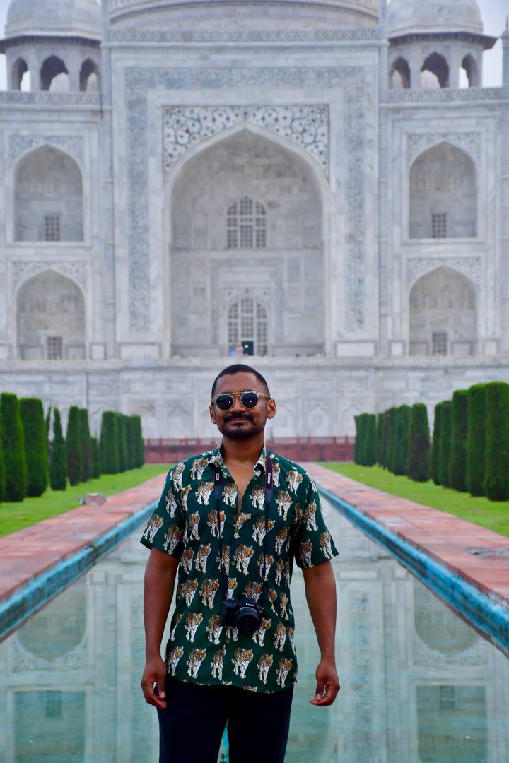 Visitor poses with sunglasses before the marble façade of the Taj Mahal, framed by gardens and cypress trees.