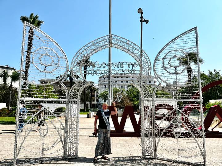 Traveller waving under a decorative metal archway that reads 'Welcome to Morocco' in a sunny plaza.