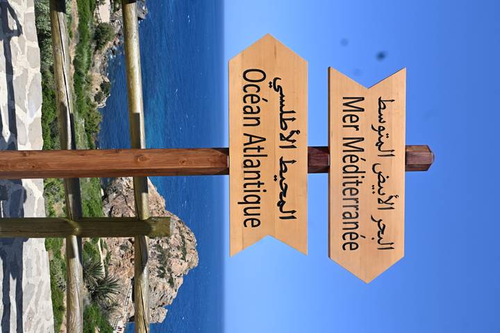 Wooden signpost pointing to the Mediterranean Sea and Atlantic Ocean with rocky coast beyond.