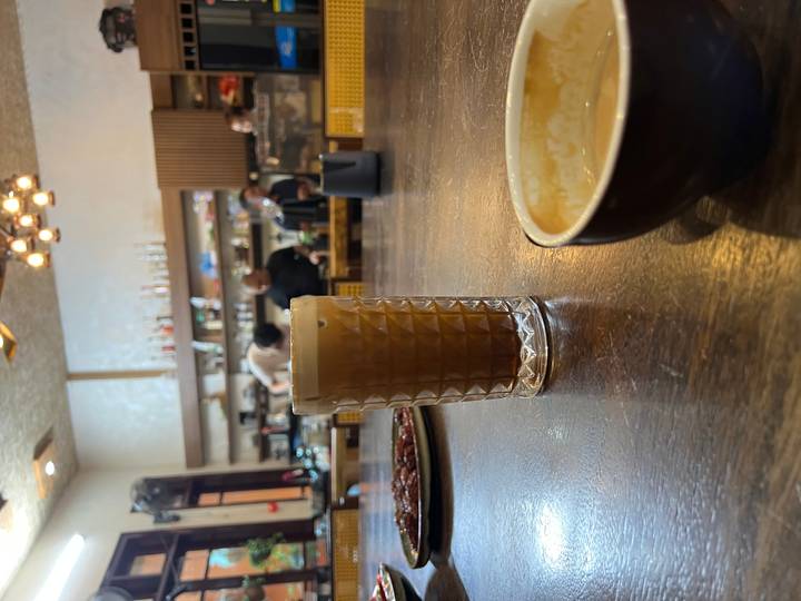 Glass of frothy Vietnamese coffee on a wooden café table with baristas working in the background.