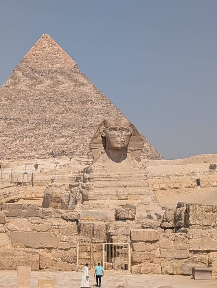 The Great Sphinx faces forward with the Pyramid of Khafre rising behind against a pale blue sky.