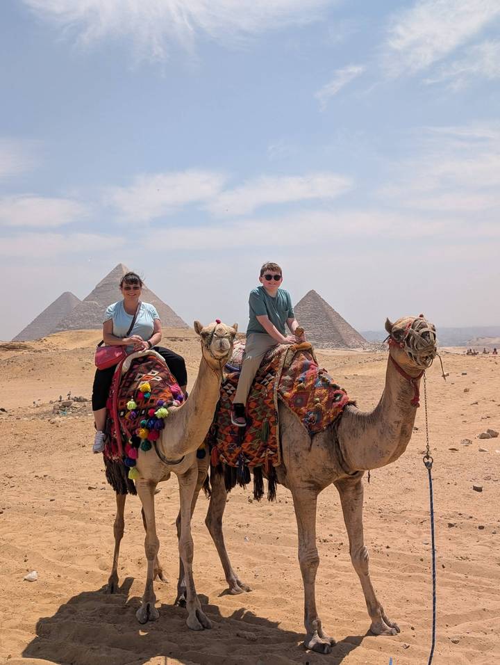 Mother and son sit atop decorated camels with the pyramids looming in the desert background.