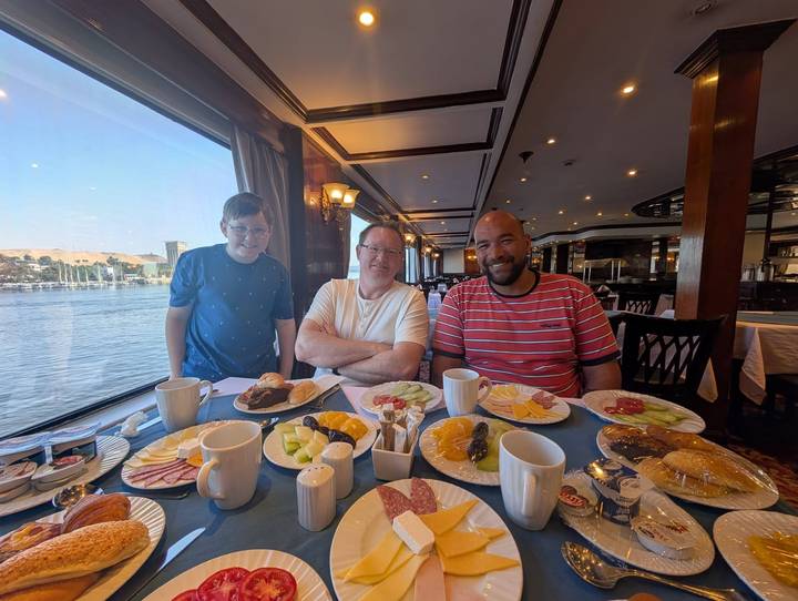 Three travelers enjoy a hearty breakfast spread on a Nile cruise ship with river views through large windows.
