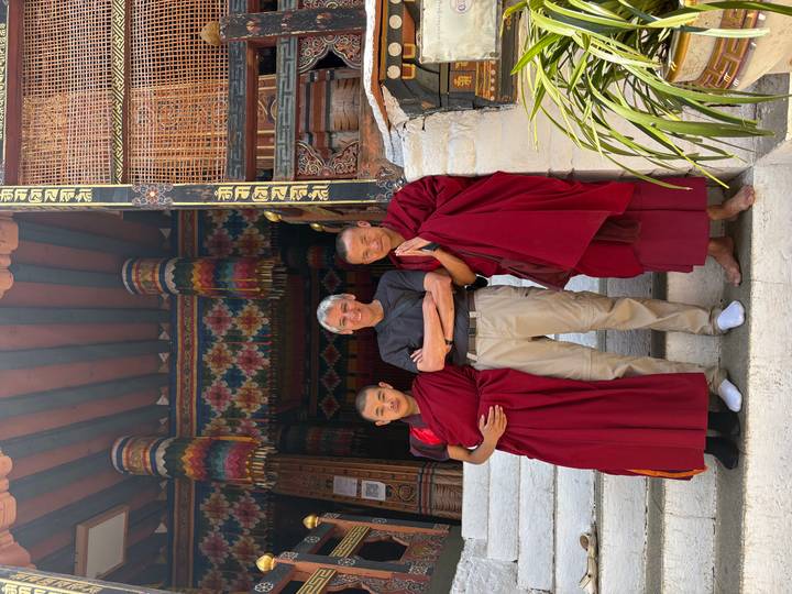 A traveler poses arm-in-arm with two monks in crimson robes on monastery steps decorated with colorful woodwork.