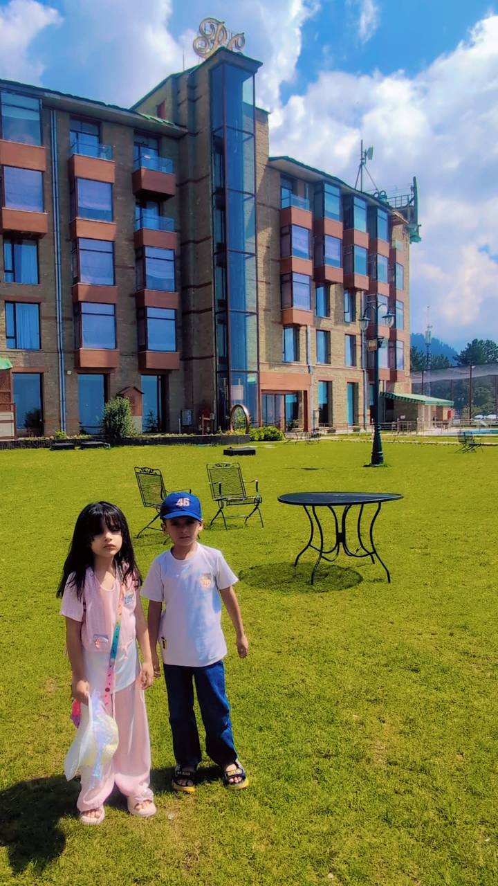 Two children stand on a manicured hotel lawn with a glass-fronted building towering behind.
