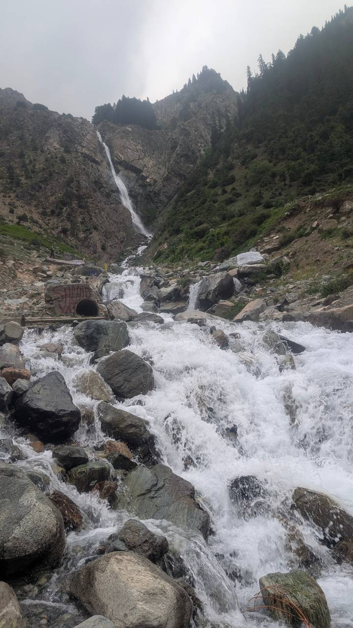 White water tumbles over boulders in a steep mountain ravine with a distant waterfall.