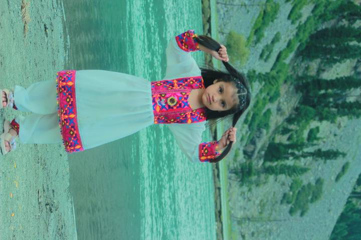 Young girl in colorful dress playfully lifts her braids beside a turquoise alpine lake.
