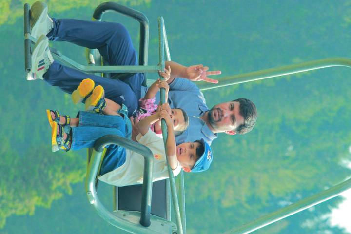 Father and two children ride an open chairlift above green forest, father flashing a peace sign.