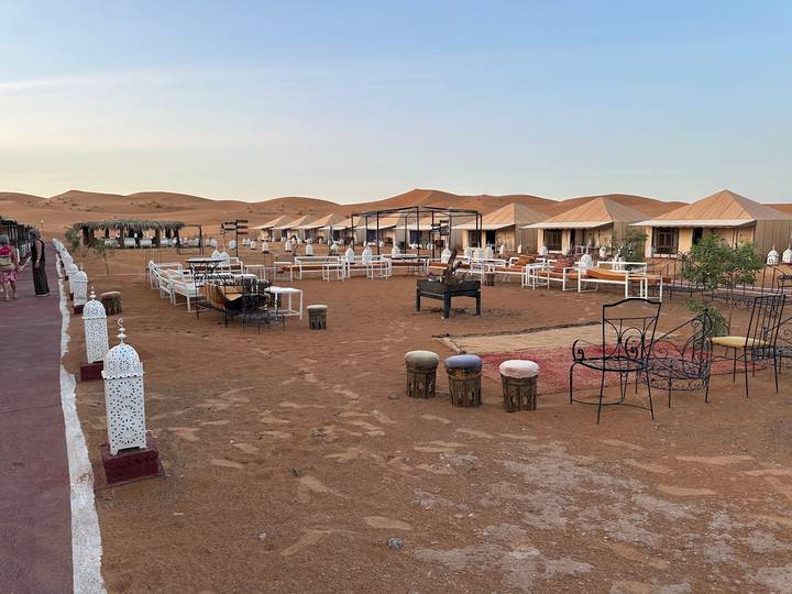 A desert camp with canvas tents, metal lanterns, and fire pits sits on reddish sand dunes at dusk.