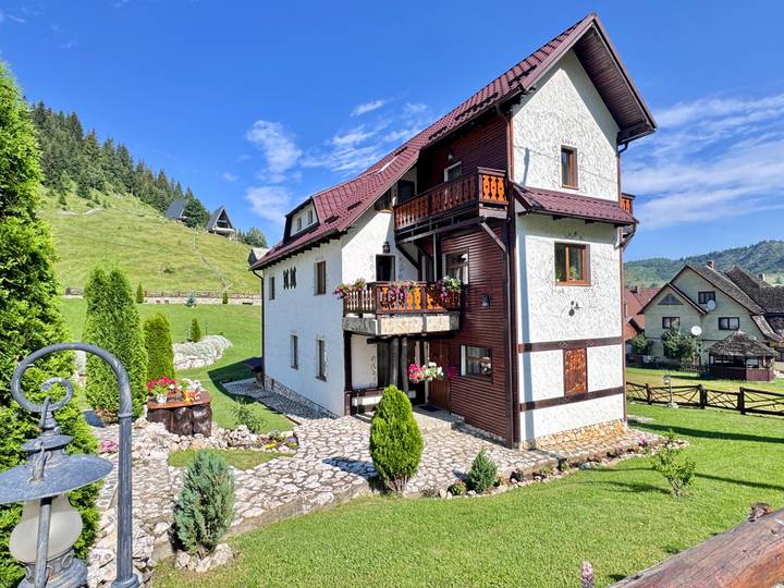 Charming alpine guesthouse with balconies of flowers set on a grassy hillside under clear skies.