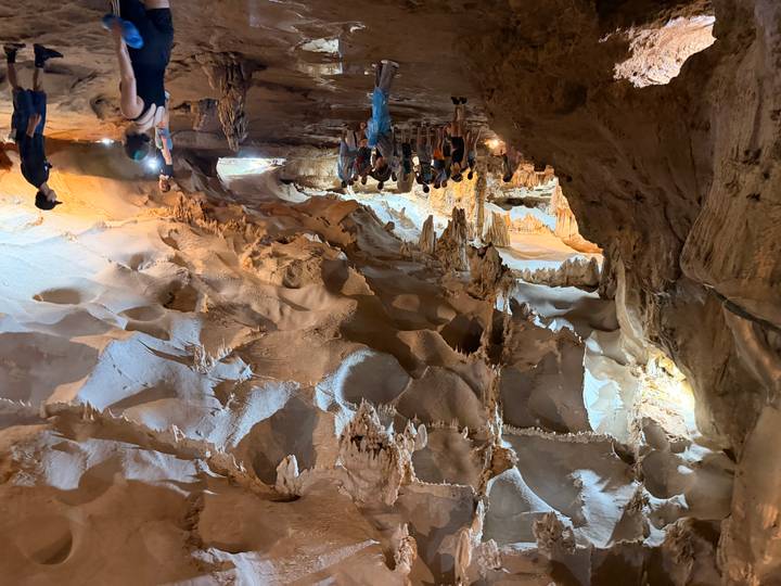 Tourists explore a vast limestone cave chamber adorned with stalactites and warm lighting.