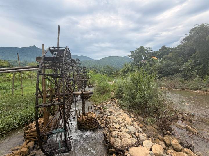 Traditional bamboo water wheels line a small rocky stream with green mountains in the background under a cloudy sky.