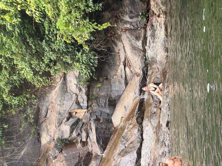 A woman in swimwear relaxes on wet rocks beside a calm natural pool at the base of a forested cliff.