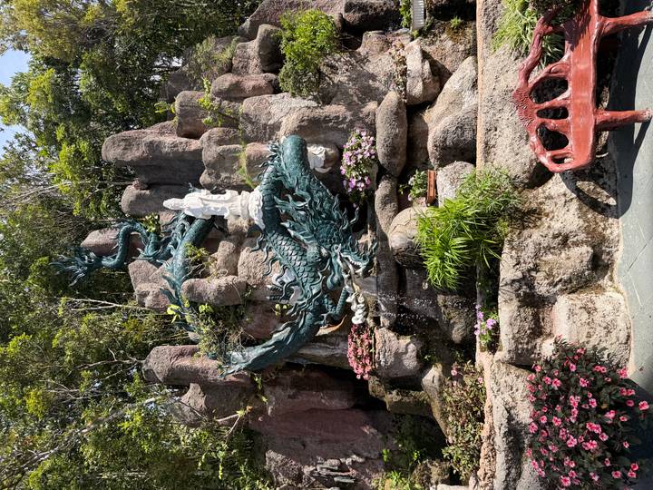 An elaborate blue-green dragon sculpture curls around rocks with a white deity statue above, surrounded by flowers and greenery.