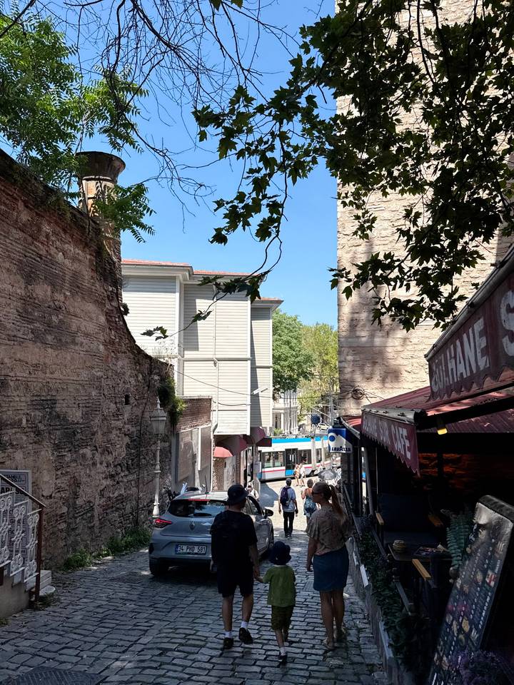 Narrow urban lane with stone walls, leafy branches and passing tram below