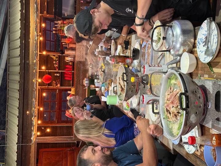 Large group enjoying steaming communal hotpot meal at a rustic outdoor restaurant strung with lights