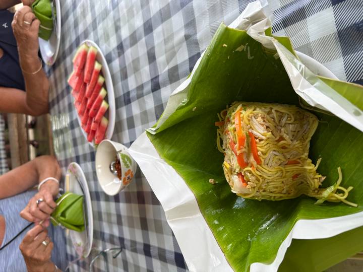 Portion of fried noodles wrapped in banana leaf with sliced watermelon in background on a checkered table
