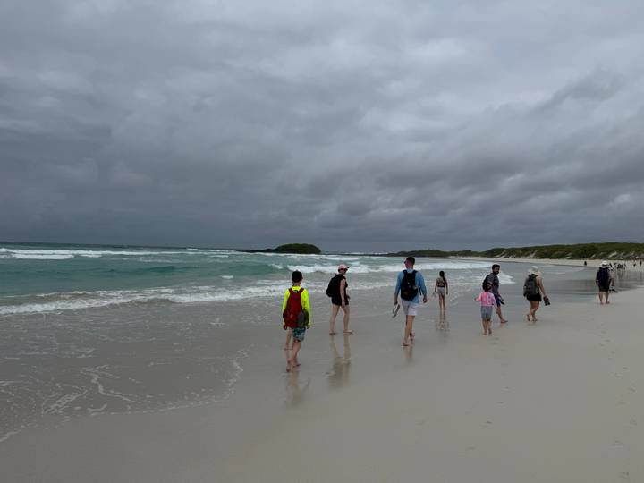 Visitors walking along wide white-sand beach with surf under gray skies