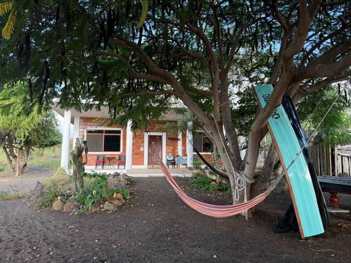 Simple island guesthouse with hammock under shady tree and surfboard resting against trunk