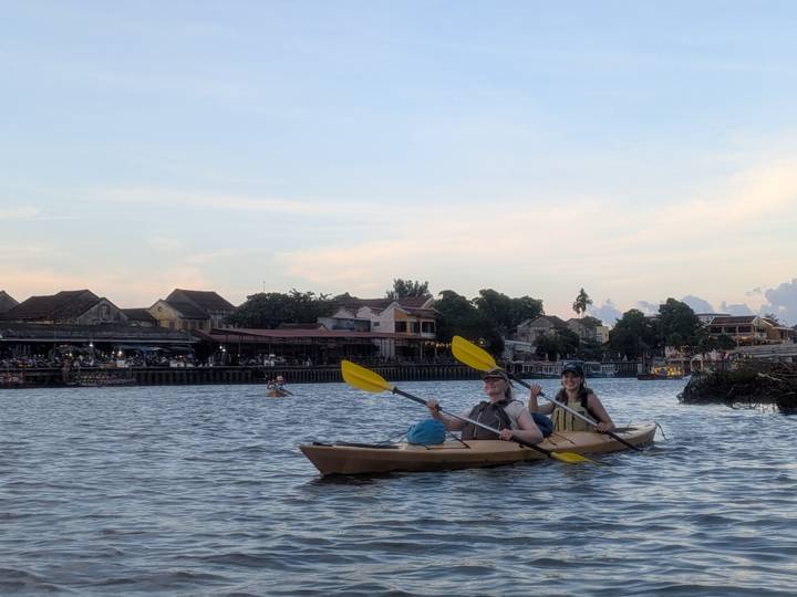 Couple paddling a tandem kayak on a calm river with Hoi An old town buildings at dusk