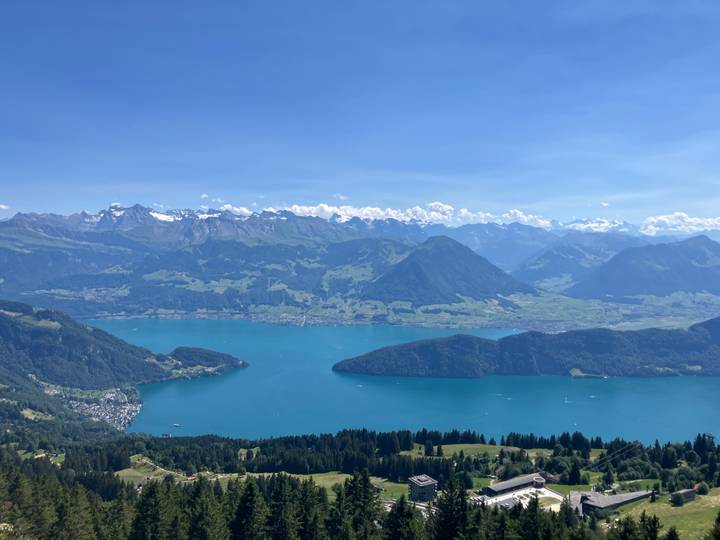 Panoramic high-altitude view of a turquoise lake encircled by green valleys and snow-dusted Alps