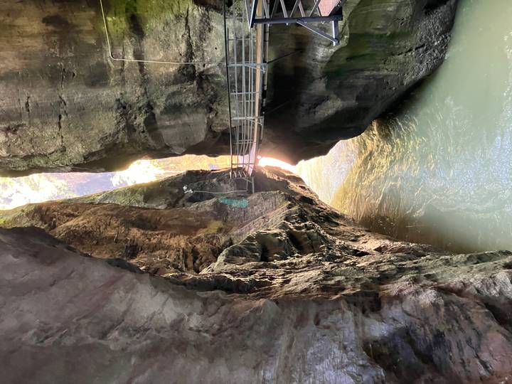 Narrow gorge with a suspended metal walkway above milky glacial water, sunlight filtering through