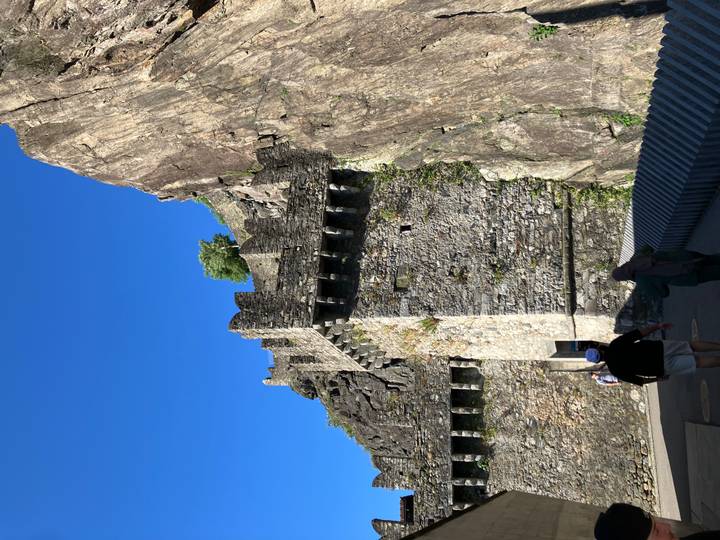 Visitors walk below the high stone walls of a medieval fortress built against a towering cliff face
