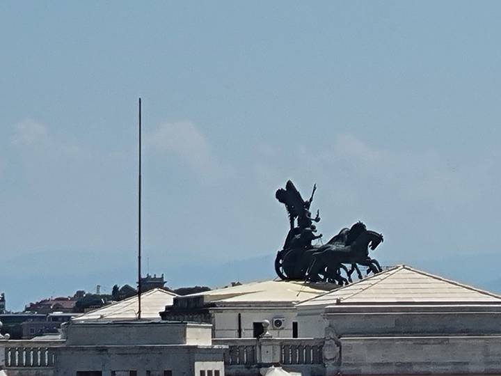 Silhouette of a chariot statue atop a classical building roof against a bright midday sky in Rome.
