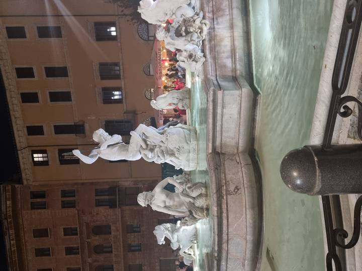 Night-lit marble statues in a Roman fountain surrounded by glowing windowed buildings.