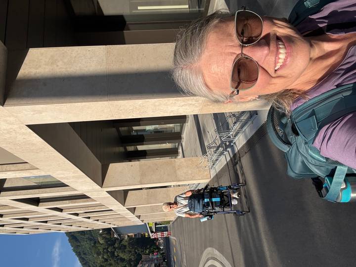 Two women, one pushing a mobility aid, enjoy sunny city streets beside a modern stone building.