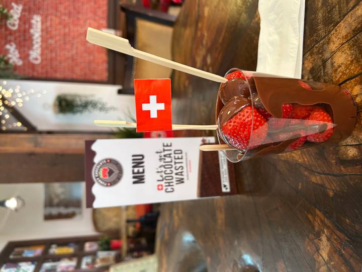 Cup of fresh strawberries dipped in melted chocolate with a small Swiss flag on top, set on a wooden café table.