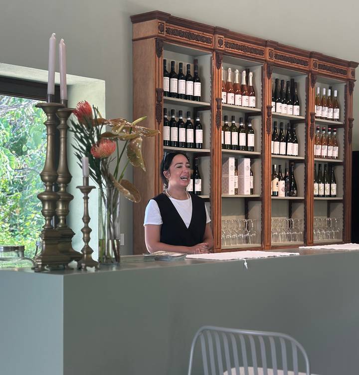 Smiling hostess stands behind a wine bar lined with bottles and glasses.