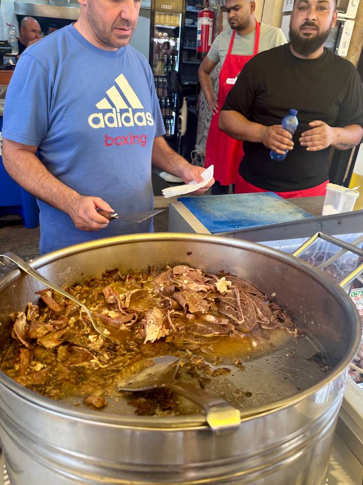 Street-food vendor slices simmering meat in a large steel pan at an open stall.