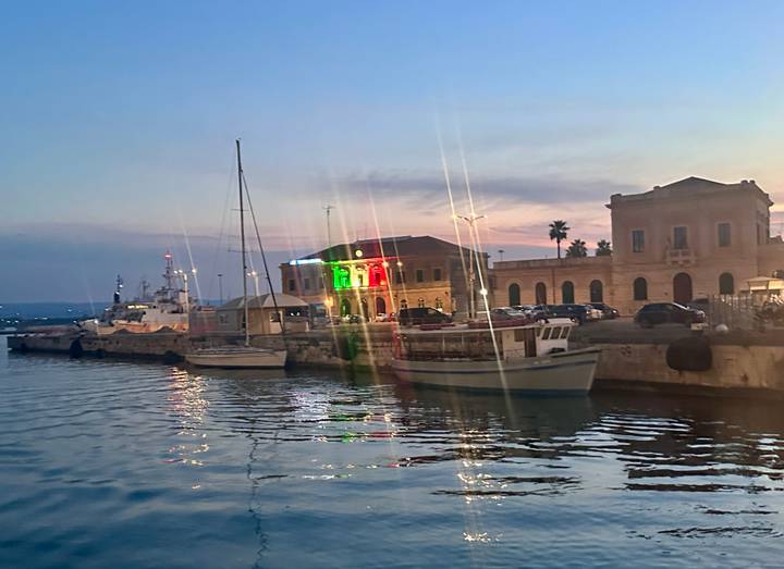 Harbor scene at dusk with boats moored beside historic buildings lit by colorful lights.
