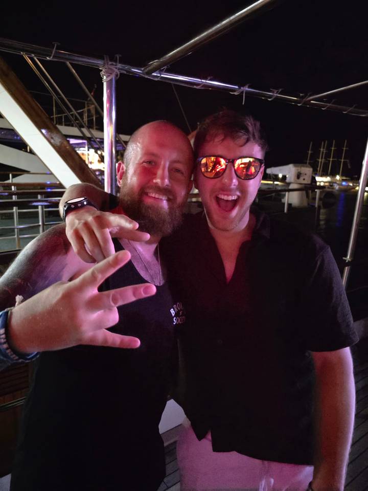 Two friends smile for a night-time party photo on a boat deck, neon reflections in sunglasses.