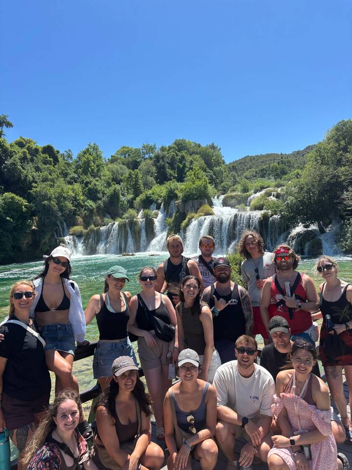Large tour group poses happily in front of cascading waterfalls and emerald water on a sunny day.