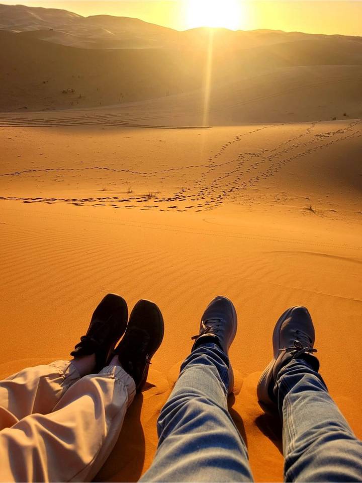 View from above of golden sand with footprints, framed by three pairs of resting feet.