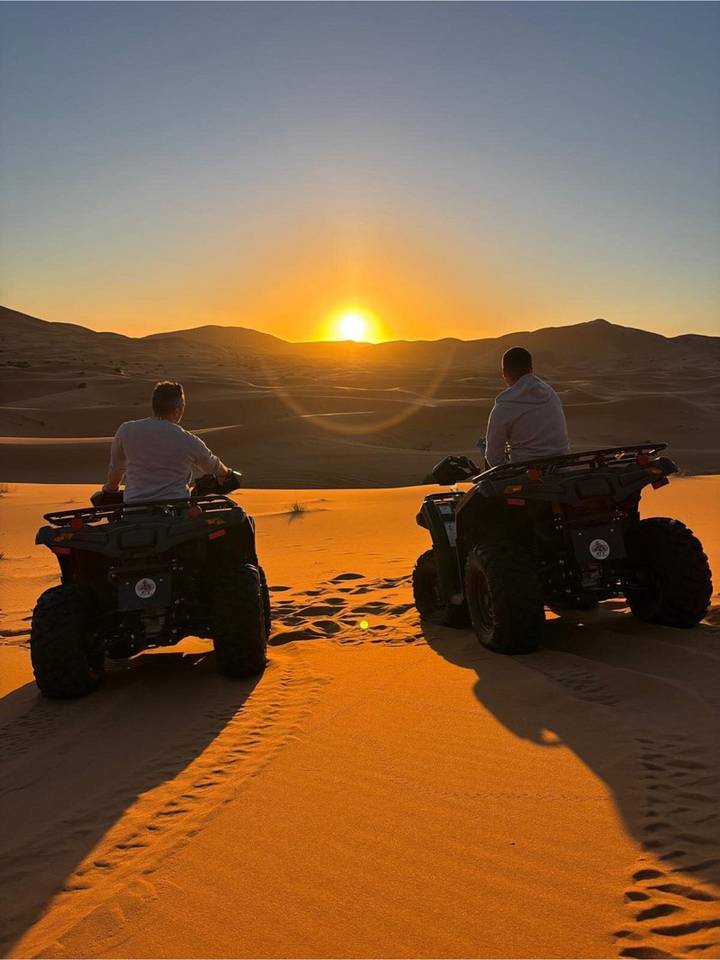 Two riders on quad bikes pause on desert sand to admire a glowing sunset.