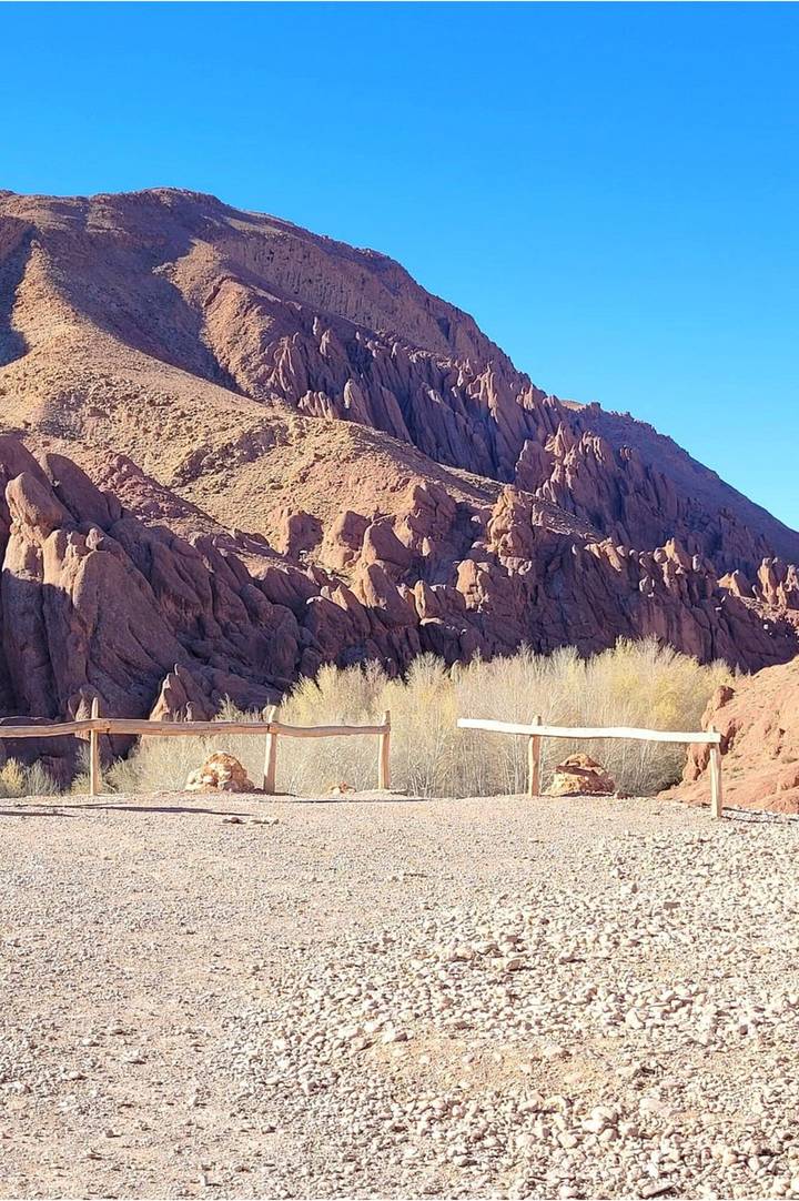 Sun-lit red rock formations with jagged shapes rising above sparse desert vegetation and a rustic wooden fence in the foreground.