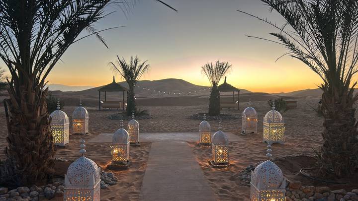 Desert camp pathway lit by ornate lanterns leading toward gazebos at twilight with sand dunes silhouetted.